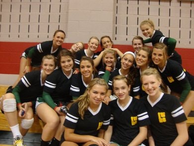 A smiling high school girls' volleyball team in black and green uniforms posing together on a gym bench.