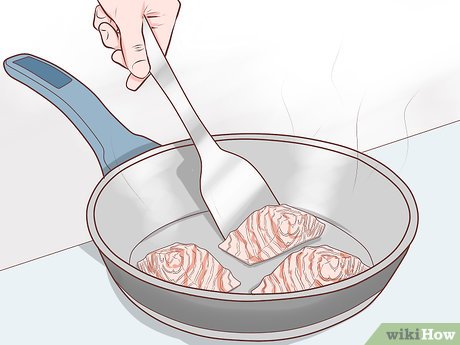 A hand uses a metal spatula to flip three raw salmon fillets sizzling in a nonstick frying pan.