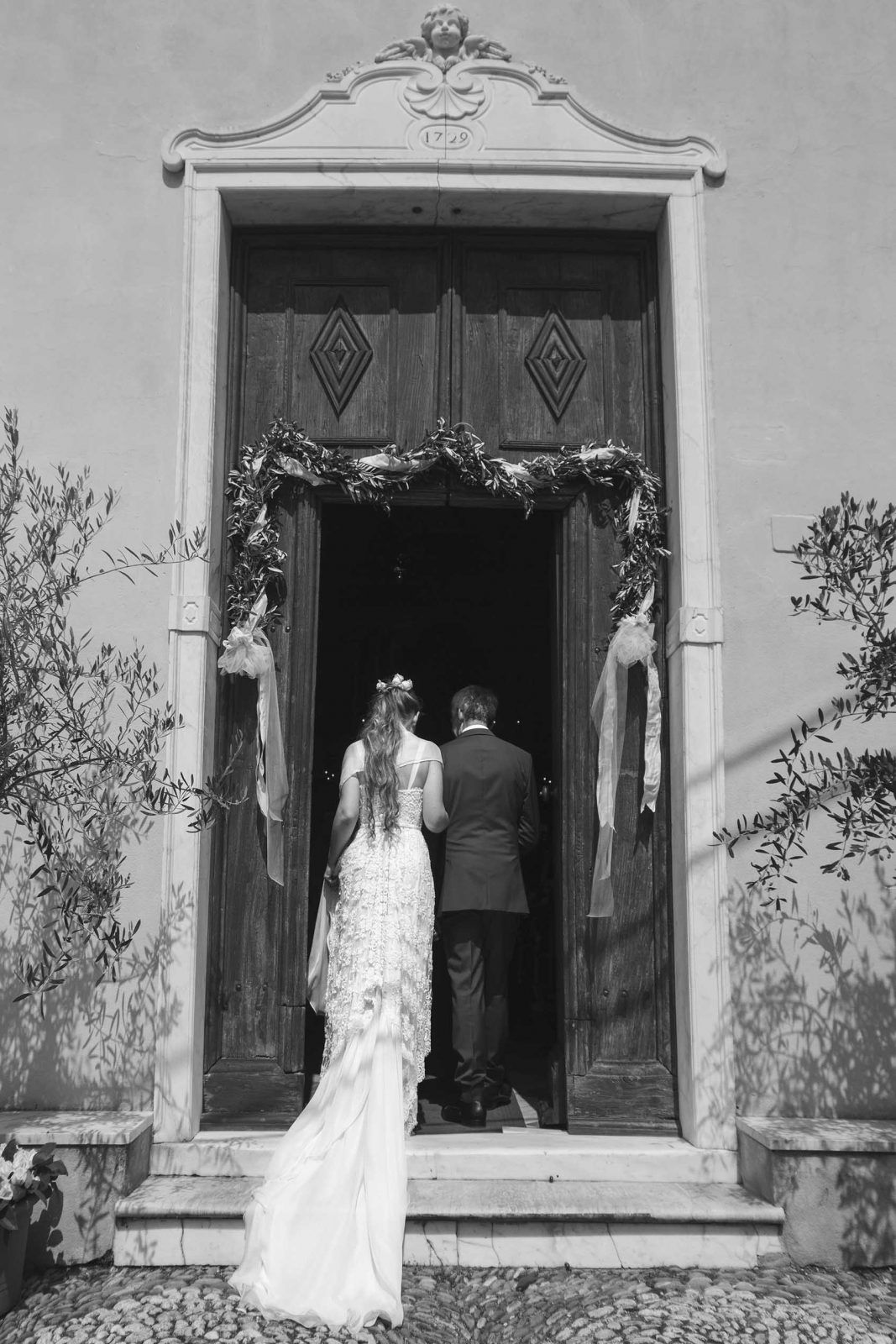 Bride and groom entering a historic church doorway adorned with olive wreaths, black-and-white wedding photo.