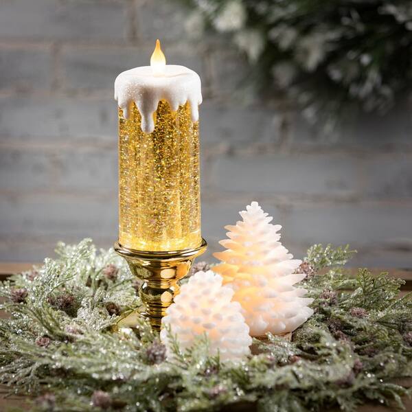 Glittering gold LED candle with faux wax drip, set on brass holder amid frosted pine trees and greenery wreath.