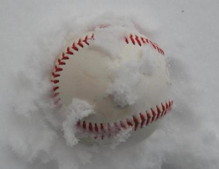 A snow-covered baseball rests in a fresh layer of white snow, red stitching partially visible.