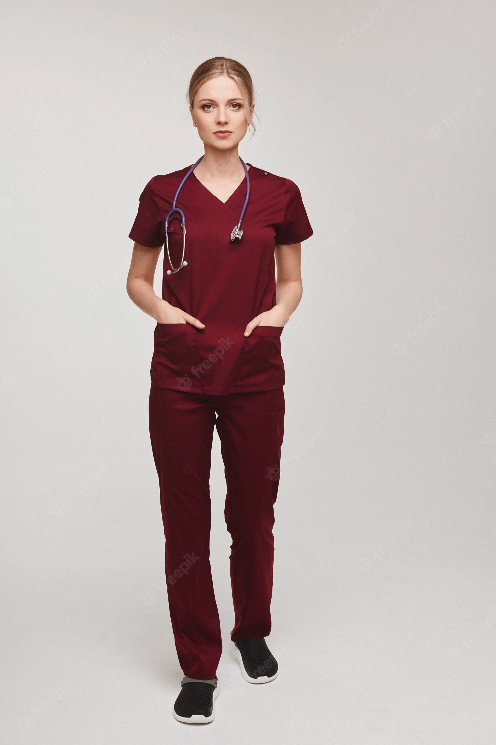 Female healthcare professional in maroon scrubs with stethoscope, hands in pockets, standing against neutral background.