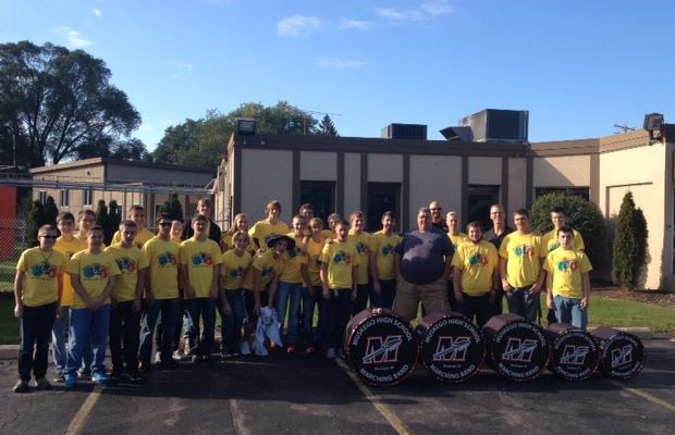 A group of students and adults in yellow shirts pose with marching band drums outside a school building on a sunny day.