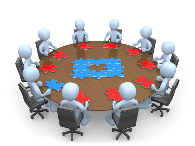 Eight stylized figures collaborate around a round table assembling red and blue puzzle pieces into a cohesive solution.