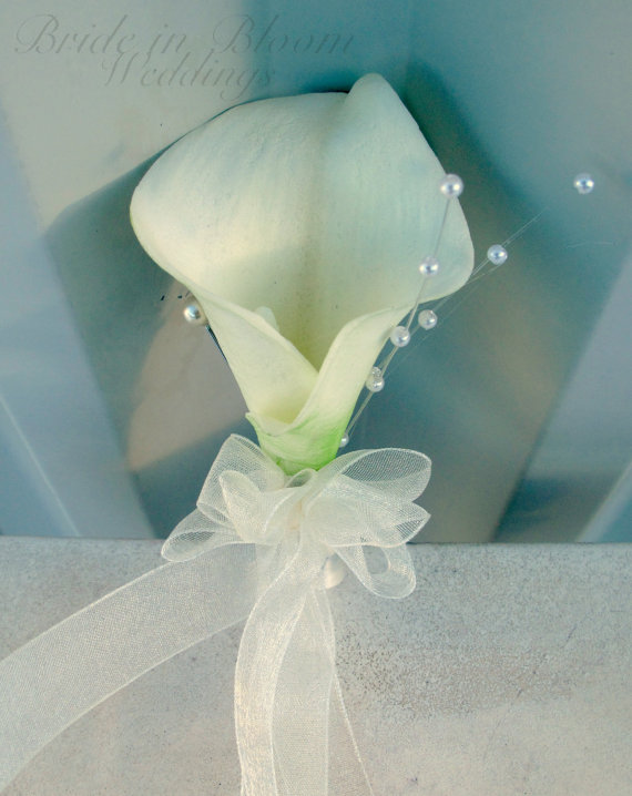 Elegant white calla lily boutonniere with pearl accents and sheer ribbon bow, set against soft blue background.
