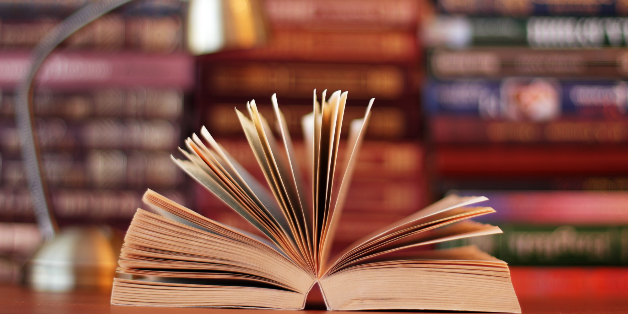 An open book with fanned pages on a wooden desk, blurred bookshelves and a desk lamp in the background.