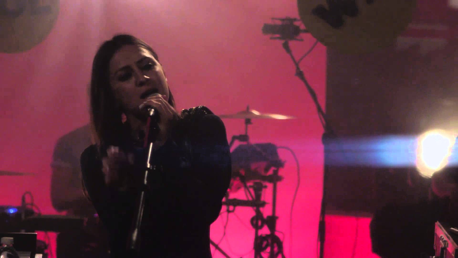 Female singer performing on stage under pink lighting with microphone and drum kit in background