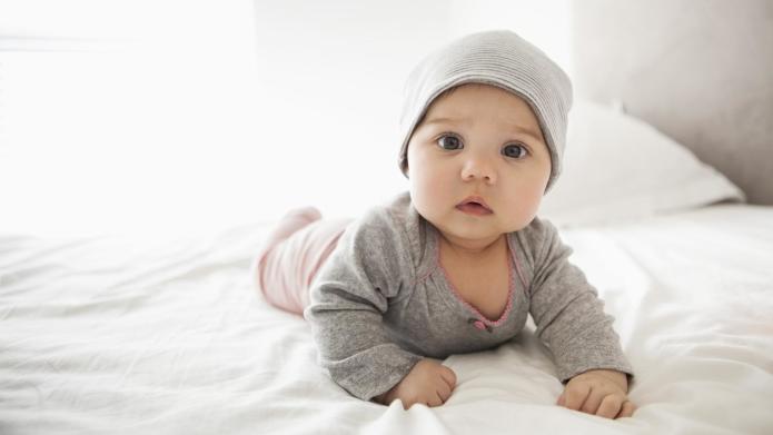 Adorable baby in gray beanie and outfit lying on white bedsheet, looking curiously at camera.