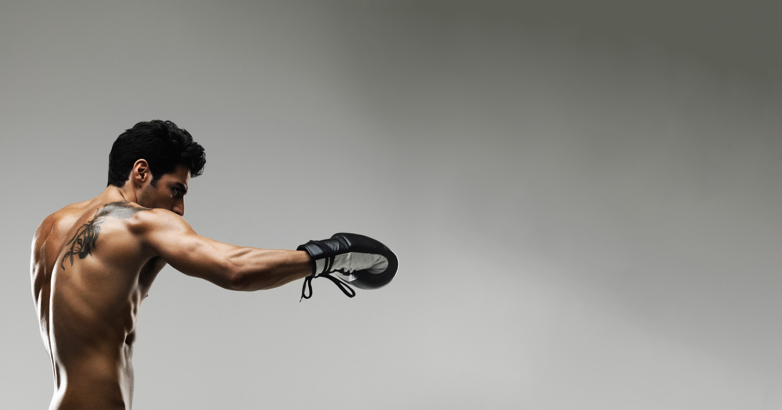 Muscular man with tattoo throwing a focused boxing punch in black gloves against gray studio background.