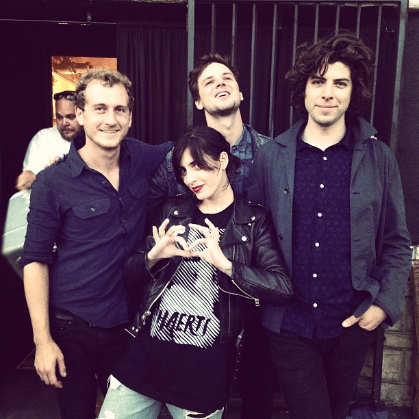 Four band members posing together backstage, one making a heart gesture with hands.