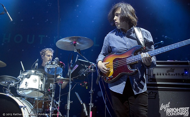 Bassist performing on stage with drummer in background under blue stage lights at a live concert.