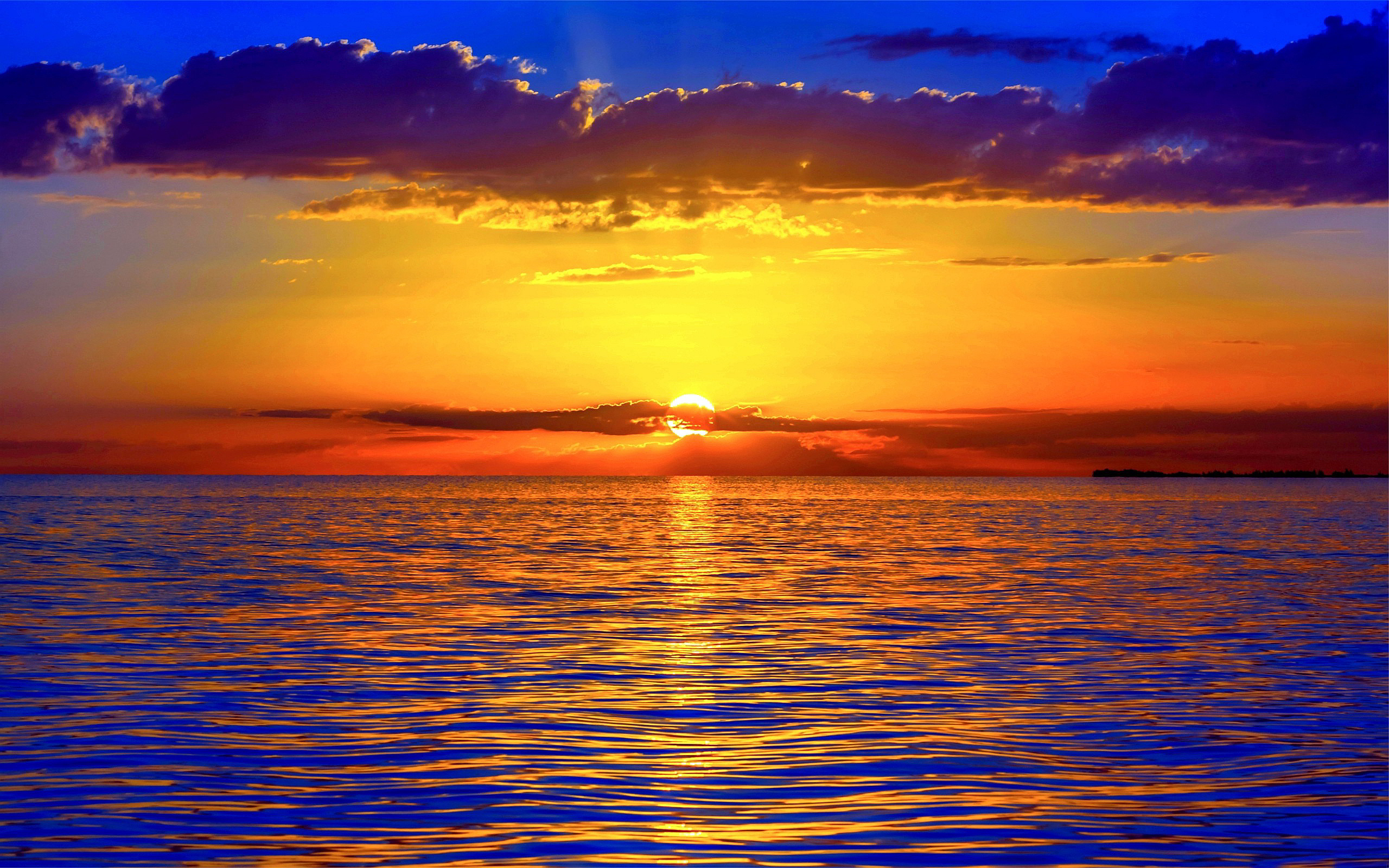 Golden sunset over calm ocean with vibrant orange, yellow, and deep blue hues reflecting on rippling water and dramatic clouds.