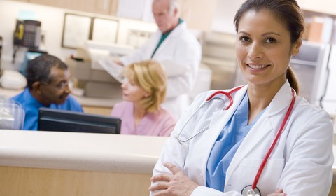 Smiling female doctor in white coat with stethoscope, standing confidently in a busy medical office setting.