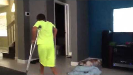 Person in bright yellow dress using crutches walks through living room while cat rests on blanket nearby.