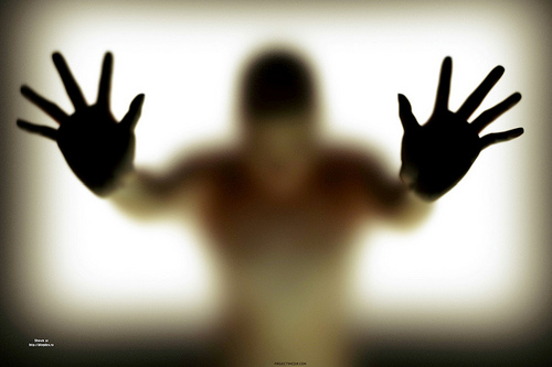 Silhouette of a person pressing hands against frosted glass, blurred figure behind translucent surface.