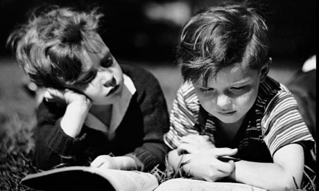 Two children lying on grass, deeply focused on reading an open book together in black-and-white.