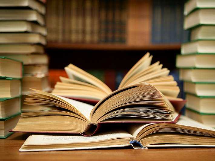 Open books fanned out on a wooden table with stacks of books and shelves in the background, evoking a scholarly library atmosphere.