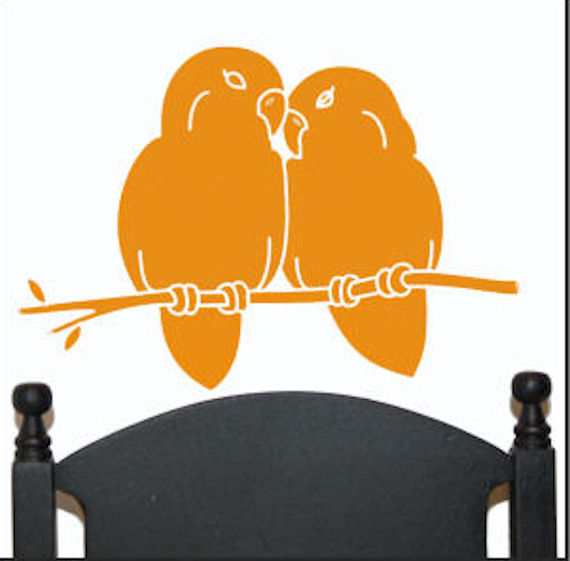 Two orange lovebirds perched on a branch, beaks touching affectionately, above a dark wooden headboard.