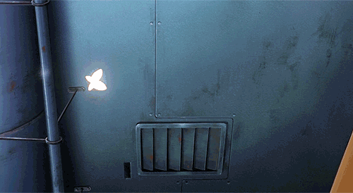 A glowing white butterfly-shaped light mounted on a weathered blue metal wall beside a vent and pipe.