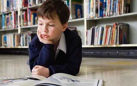 A thoughtful schoolboy in uniform sits on the library floor, resting his chin on his hand while reading an open book.