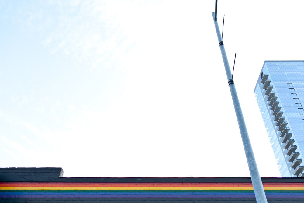 Rainbow-painted brick wall beneath a modern glass skyscraper and utility pole, symbolizing urban LGBTQ+ pride.