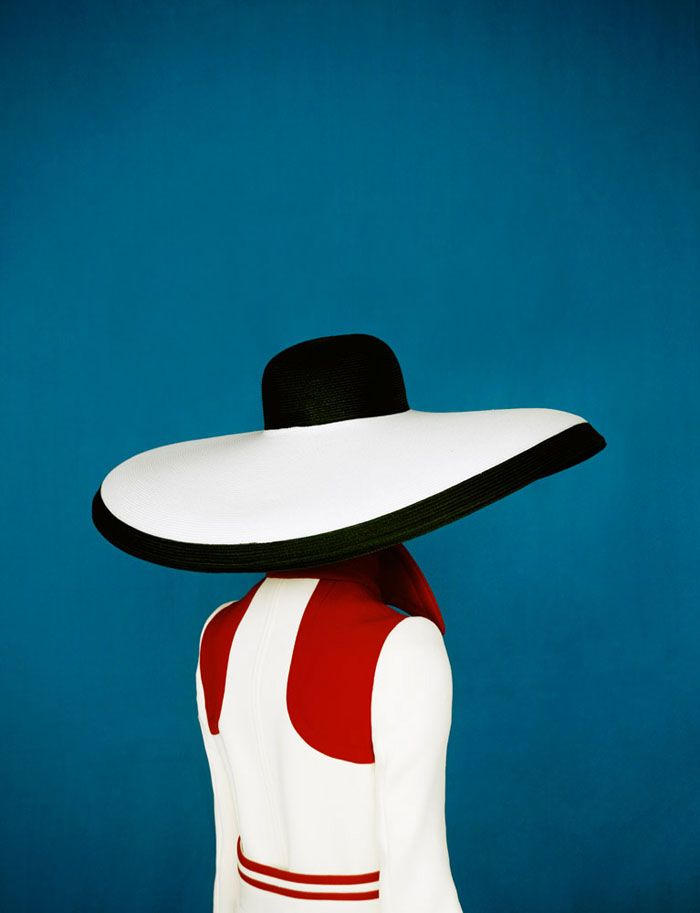 Back view of a person wearing a dramatic white-and-black wide-brimmed hat and a red-and-white sleeveless outfit against a deep blue background.