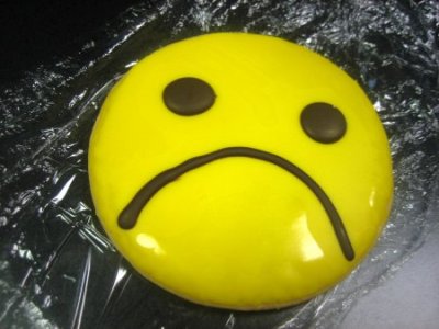 A glossy yellow sad face emoji cake with chocolate eyes and frown, resting on crinkled plastic wrap.