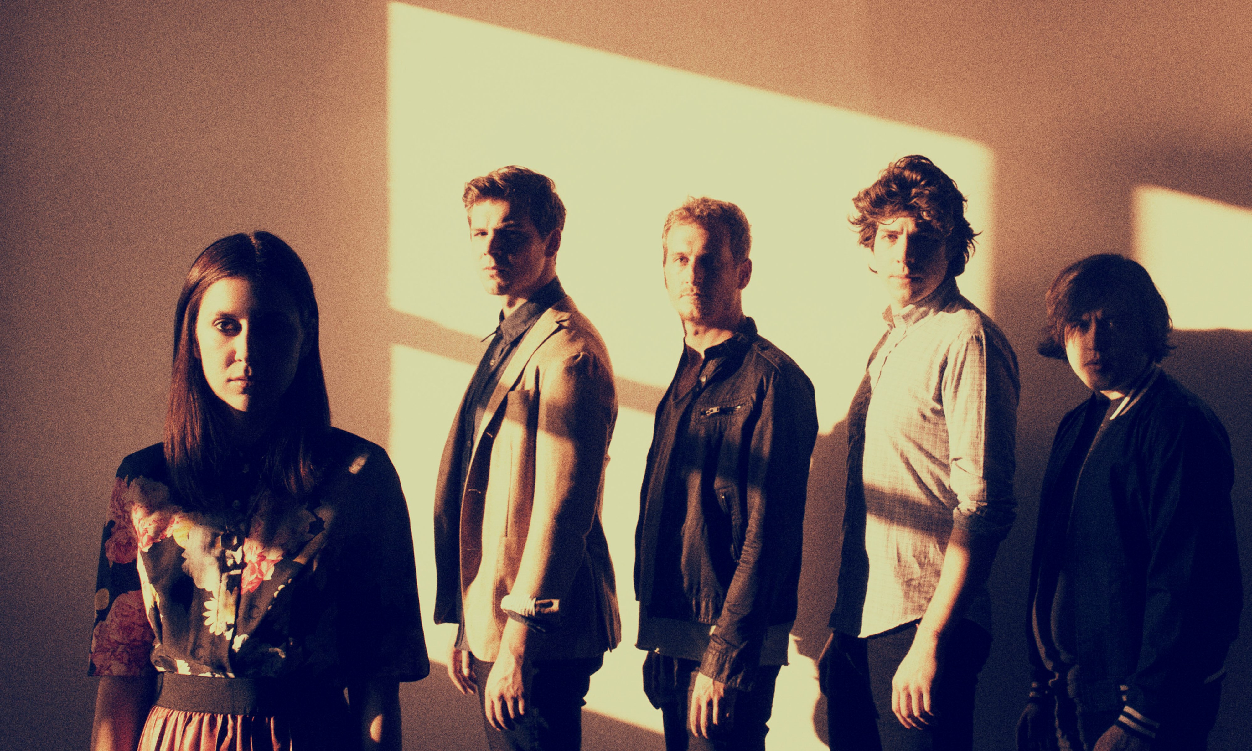 Five band members standing in dramatic chiaroscuro lighting, facing the camera with serious expressions.