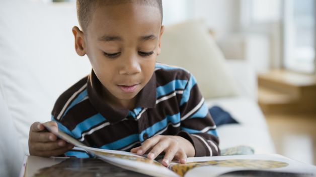 Young Black boy in striped shirt reading a picture book on a white couch.