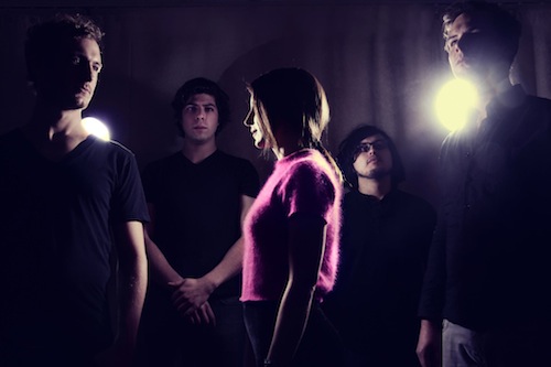 Five musicians in dramatic lighting, one woman in pink sweater center stage, others in shadows with spotlights.