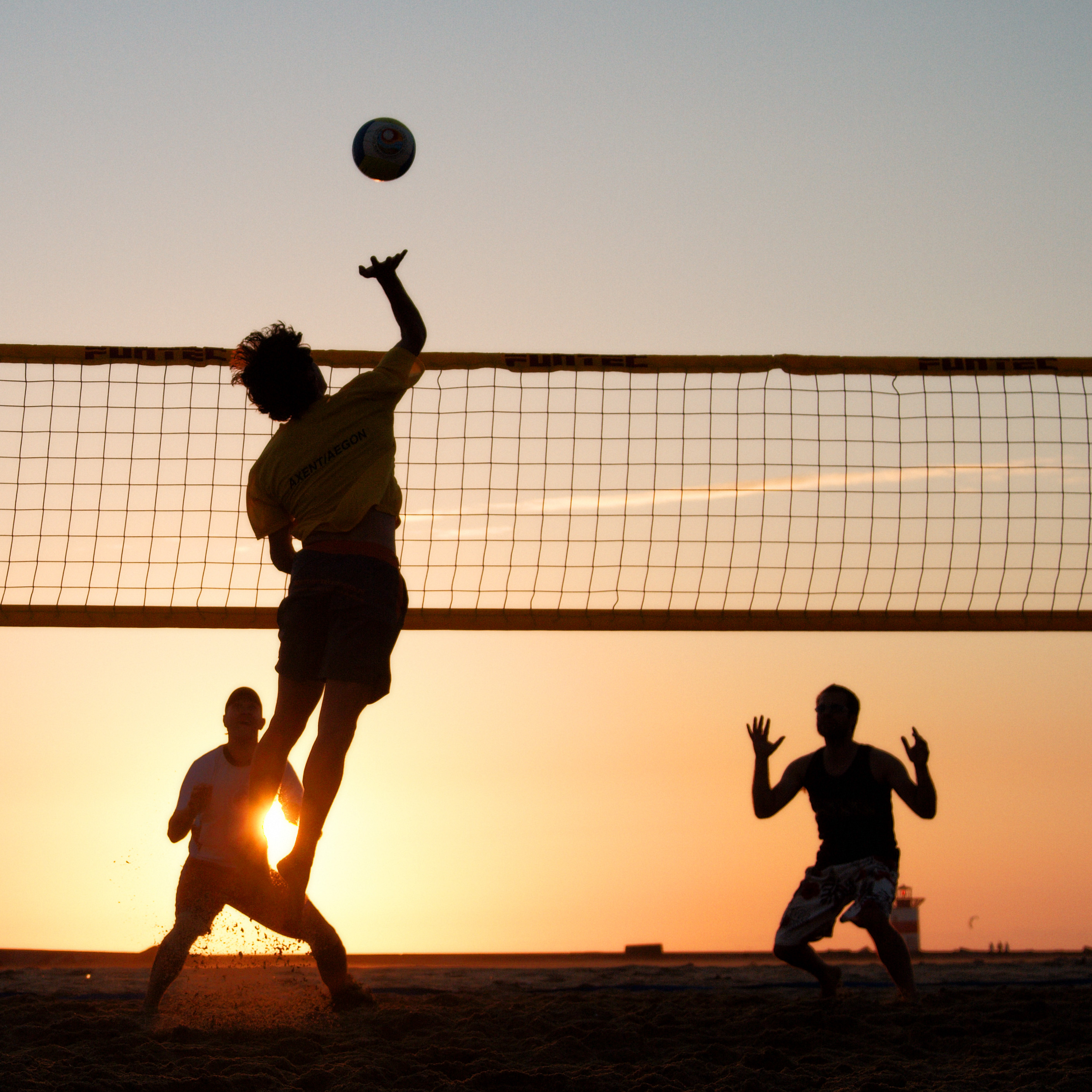 Silhouetted beach volleyball players in action at sunset, one spiking the ball over the net while teammates react.