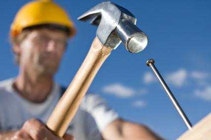 Construction worker in yellow hard hat holding a claw hammer poised to drive a nail into wood against a blue sky.