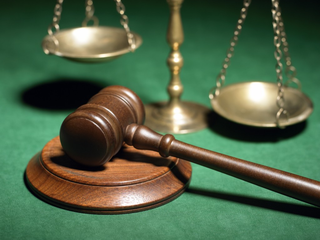 Wooden judge's gavel resting on sound block beside brass scales of justice on green felt background.