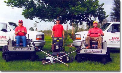Three lawn care workers in red shirts posing with commercial mowers and Eden Lawn Care trucks on a grassy field.