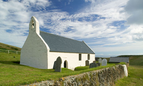 A small white stone chapel with a slate roof and bell gable, surrounded by grassy hills and old gravestones under a partly cloudy sky.