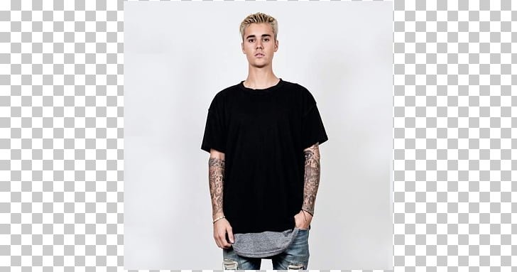 Justin Bieber wearing a black t-shirt and jeans with tattoos on his arms, standing against a white background.
